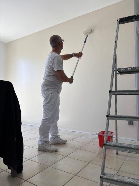 Un homme en vêtements blancs peint un mur avec un rouleau depuis une échelle.